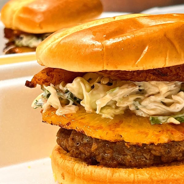 Close-up of a Jamaican burger with pineapple, coleslaw, and a crispy plantain on a bun.